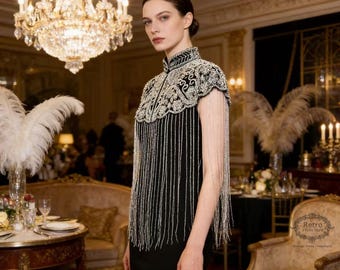May include: A black dress with a high collar and beaded embellishments. The dress features a decorative beaded capelet with long fringe. The model is standing in a room with a chandelier and a vintage style chair.