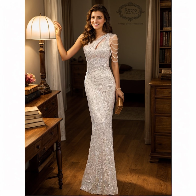 May include: A woman wearing a silver sequined one-shoulder gown with a beaded strap. The gown has a fitted bodice and a flowing skirt. The woman is holding a small gold clutch.