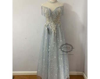 May include: A light blue tulle gown with a beaded bodice and a star print. The gown has a sweetheart neckline and off-the-shoulder straps with fringe details. The gown is on a mannequin.
