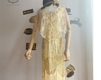 May include: A gold sequined flapper dress with fringe detailing. The dress has a V-neckline and short sleeves. The dress is on a mannequin with a black hat and feather.