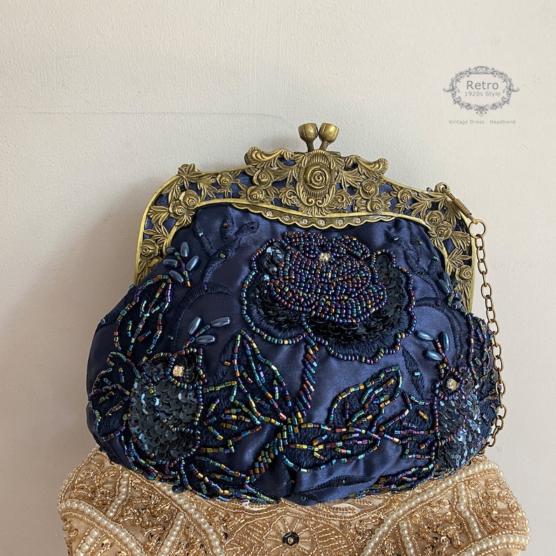 Pu&ograve; includere: Una borsa da sera in satin blu navy con una cornice in metallo dorato e una tracolla a catena. La borsa &egrave; decorata con paillettes e perline iridescenti in un motivo floreale.