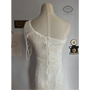May include: A white beaded one-shoulder dress with a long beaded strap on the left shoulder. The dress has a fitted silhouette and a zipper closure in the back. The dress is on a white mannequin.