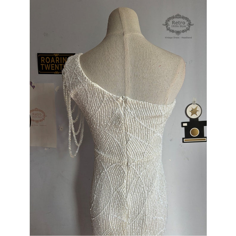 May include: A white beaded one-shoulder dress with a long beaded strap on the left shoulder. The dress has a fitted silhouette and a zipper closure in the back. The dress is on a white mannequin.