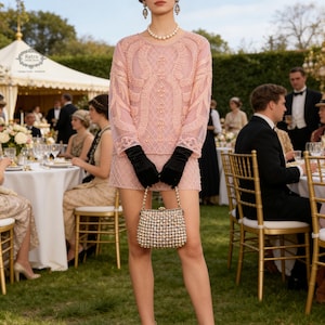 Puede incluir: Una mujer con un vestido rosa con cuentas, guantes negros y un bolso con cuentas está de pie al aire libre. Lleva un collar de perlas, pendientes y una diadema con plumas. La escena sugiere un evento formal vintage con invitados sentados a la mesa.
