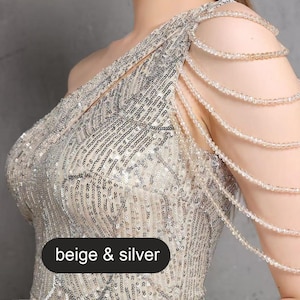 May include: A one-shoulder sequined dress with a beige and silver color scheme. The dress features a beaded strap detail on one shoulder.