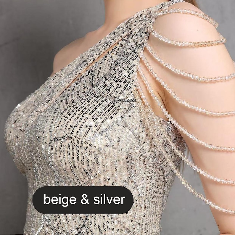 May include: A one-shoulder sequined dress with a beige and silver color scheme. The dress features a beaded strap detail on one shoulder.