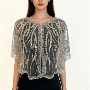Peut inclure: Cape argenté à paillettes avec un décolleté en V. Le tissu transparent est orné de perles et de paillettes argentées complexes, créant un effet scintillant. La cape a une fermeture boutonnée sur le devant et des manches courtes. Le vêtement est porté sur un haut noir.