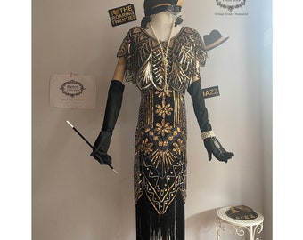 Peut inclure: Une robe de flapper noire et dorée avec des franges et des embellissements perlés. La robe est sur un mannequin et a une longue jupe fluide. La robe est stylisée avec un chapeau noir, des gants et un collier de perles. Le texte "I ♥ THE ROARING TWENTIES" est visible en arrière-plan.