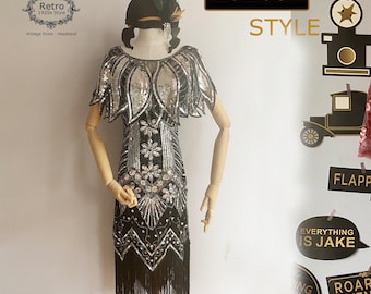 May include: A black and silver sequined flapper dress with fringe. The dress has a short sleeve and a V-neckline. The dress is on a mannequin. The image also includes props with the text "1920s Style", "Flap", "Everything is Jake", and "Roar Twenties".