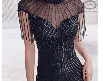 Puede incluir: Un vestido negro de lentejuelas con una superposición de malla transparente y detalles de flecos. El vestido tiene un escote alto y una silueta ajustada.