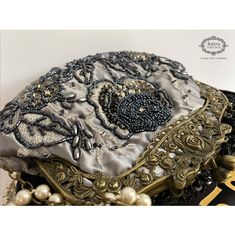 Pu&ograve; includere: Una pochette in satin argento con un motivo floreale perlato e una cornice in metallo dorato. La pochette ha una tracolla a catena e un'applicazione di perle.