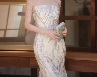 May include: Elegant ivory-colored formal gown with silver sequin and bead embellishments. The dress features a high neckline, sheer shoulder detailing, and a mermaid silhouette. A silver clutch is held in the hands.