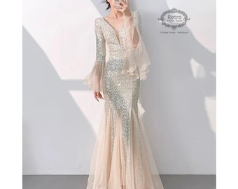 Puede incluir: Un vestido de noche largo de manga larga, con lentejuelas, estilo sirena, con escote en V y mangas acampanadas. El vestido es de color champán claro con lentejuelas plateadas. El vestido tiene una superposición de tul que crea un look fluido y etéreo. La modelo lleva el vestido con un peinado sencillo y un maquillaje mínimo.