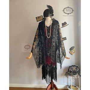 Può includere: Un scialle nero con paillettes e frange con un motivo floreale. Lo scialle è drappeggiato su un manichino che indossa un abito rosso con perline e un cappello nero con una piuma. Il testo "I ♥ THE ROARING TWENTIES" è visibile sullo sfondo.