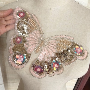 Puede incluir: Aplique decorativo de mariposa grande con lentejuelas y cuentas rosas, doradas y marrones. La mariposa tiene un diseño delicado y en capas con detalles florales y está adherida a una malla transparente.