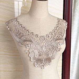 Może przedstawiać: Beżowa aplikacja koronkowa, z kwiatowym wzorem pośrodku i falistym brzegiem. Aplikacja jest prezentowana na manekinie, podkreślając jej skomplikowane detale i delikatne rzemiosło. Koronka jest w neutralnym kolorze.