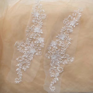 May include: Two white floral lace appliques with intricate designs. The appliques feature detailed flower and leaf patterns, perfect for embellishing garments or crafts. The lace is set against a light beige background.