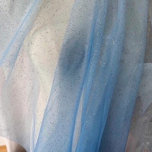 May include: A sheer, light blue fabric with a gradient effect, transitioning from a lighter shade to a deeper blue. The fabric is sprinkled with tiny, sparkling glitter particles, creating a shimmering effect. Ideal for crafting or decorative purposes.