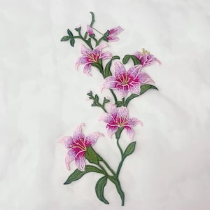 May include: Embroidered floral applique featuring pink and white lilies with green leaves and stems. The flowers have a detailed design with a dark pink centre and light pink edges. The applique is on a white background.