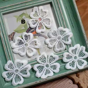 Könnte beinhalten: Sechs weiße, durchbrochene Blumenapplikationen mit filigranem, spitzenartigem Design. Die Applikationen sind auf einem gerahmten Bild mit grünem Rand angeordnet. Die Blumen haben eine fünfteilige Form mit einem detaillierten, netzartigen Zentrum.