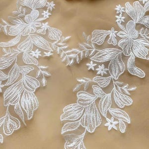 May include: Close-up of white floral lace embroidery. The design features detailed leaves, flowers, and small star-shaped accents. The lace is set against a soft, neutral background, highlighting the intricate patterns.