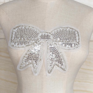 Könnte beinhalten: Silberne Pailletten-Schleifenapplikation mit weißem Hintergrund. Der Bogen ist dicht mit kleinen, glänzenden Pailletten und Perlen bedeckt, was einen funkelnden Effekt erzeugt. Die Applikation dient wahrscheinlich zum Verzieren von Kleidung oder Accessoires.