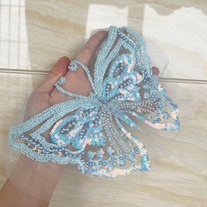 May include: A light blue butterfly applique, embellished with beads and sequins. The butterfly design features intricate details, with a delicate net backing. The wings showcase a gradient of colors, including light blue, pink, and iridescent sequins.