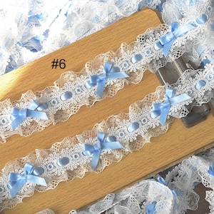 Puede incluir: Ribete de encaje blanco con borde festoneado, adornado con lazos de satén azul claro y detalles ovalados. El ribete se muestra sobre una superficie de madera. El texto "#6" es visible. Adecuado para manualidades y costura.