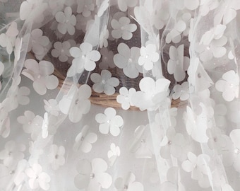 May include: Close-up of white floral fabric. The fabric features three-dimensional white flowers with small, sparkling embellishments. The flowers are scattered across a sheer, white background, creating a delicate and textured appearance. The fabric is likely used for decorative purposes.