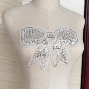 Może przedstawiać: Srebrna aplikacja w kształcie kokardy z cekinami na białym podkładzie. Kokarda jest gęsto pokryta małymi, błyszczącymi cekinami i koralikami, tworząc efekt iskrzenia. Aplikacja prawdopodobnie służy do ozdabiania odzieży lub akcesoriów.