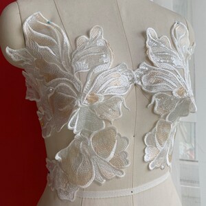 Może przedstawiać: Biała aplikacja z koronki kwiatowej o delikatnym wzorze. Koronka wykonana jest z przezroczystej tkaniny z subtelnym połyskiem. Aplikacja idealnie nadaje się do dodania odrobiny elegancji do każdego ubrania.