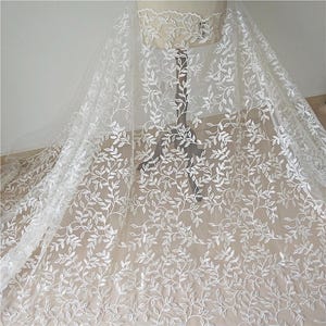 Può includere: Tessuto di pizzo bianco con un delicato motivo a foglie. Il tessuto è drappeggiato su un manichino, mostrando l'intricato ricamo floreale. Il materiale trasparente lascia passare la luce, evidenziando il design dettagliato.