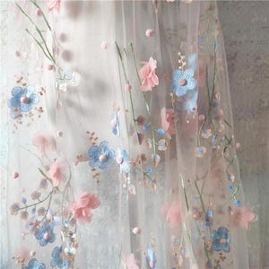 Puede incluir: Tela transparente con diseño floral bordado. La tela presenta flores azules claras y rosas con tallos verdes y pequeños detalles rosas. Las flores están dispersas sobre el material transparente y claro.