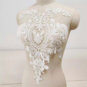 Pode incluir: Aplique de renda branca com um design floral. A renda é delicada e intrincada, com uma borda festonada. Este aplique seria perfeito para adicionar um toque de elegância a um vestido de noiva, um vestido de noiva ou outra roupa para uma ocasião especial.