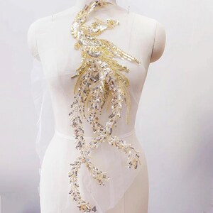 May include: A gold and silver sequined applique in a flowing, asymmetrical design. The embellishment features a central, feathered motif with cascading, leafy strands. The sequins create a shimmering effect, ideal for embellishing garments or accessories.