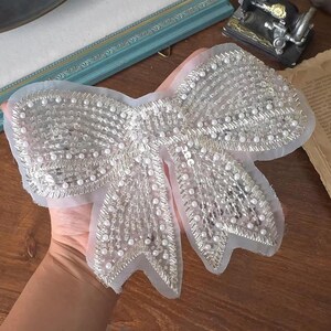 May include: A large, silver-colored bow applique, heavily embellished with beads and sequins. The bow is made of a sheer, light-colored fabric and features a detailed, textured design. The decorative element is ideal for embellishing clothing or accessories.