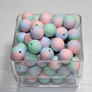 Puede incluir: Un recipiente cuadrado transparente lleno de cuentas de silicona de colores pastel. Las cuentas son redondas, con un agujero en el centro, y son una mezcla de rosa, azul y verde. Las cuentas son probablemente para manualidades o fabricación de joyas.