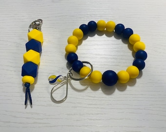 May include: A matching set of yellow and navy blue accessories. The set includes a beaded bracelet and a keychain. The keychain has a small charm with a blue and yellow ribbon. The bracelet is made of round beads, and the keychain has cube-shaped beads.