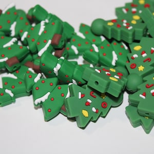 May include: A collection of green Christmas tree-shaped beads with white accents, red dots, and colorful decorations. The beads have a brown trunk and are scattered on a white surface. These beads are likely for crafting or jewelry making.