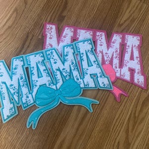May include: Three felt patches with the word "MAMA" in white letters with a floral pattern on a pink and blue background. The blue patch has a turquoise bow.