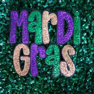 Op de afbeelding: Een Mardi Gras bord gemaakt van paarse, groene en gouden pailletten. Het bord is gemaakt van afzonderlijke letters en is geplaatst tegen een groene achtergrond.