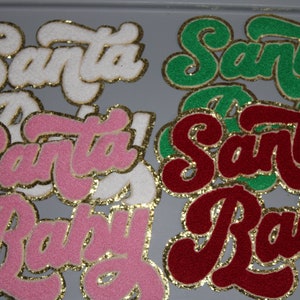 May include: Assortment of felt patches with gold glitter trim. The patches feature the words "Santa" and "Baby" in a variety of colors, including white, pink, red, and green.