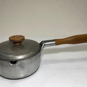 May include: A silver-coloured metal saucepan with a matching lid and a long wooden handle. The lid features a small, round wooden knob at the centre. The pan has a rounded body with a small pouring spout on the side, showing a matte finish.