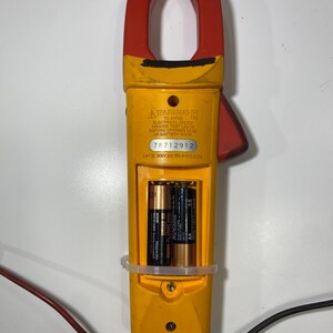 Pu&ograve; includere: Un multimetro a pinza giallo e rosso con vano batteria aperto contenente due batterie AA. Il dispositivo presenta un'etichetta di avvertenza, il numero di serie 78712912 e il testo CAT III 600V. Cavi di prova rosso e nero sono collegati.