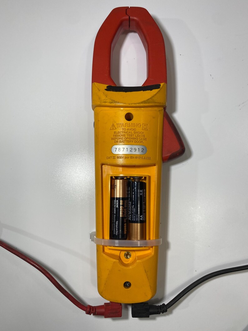 Pu&ograve; includere: Un multimetro a pinza giallo e rosso con vano batteria aperto contenente due batterie AA. Il dispositivo presenta un'etichetta di avvertenza, il numero di serie 78712912 e il testo CAT III 600V. Cavi di prova rosso e nero sono collegati.