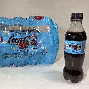 以下が含まれることがあります： ライトブルーのプラスチックフィルムで包まれた300mlのコカ・コーラ ドリームワールド ゼロシュガー8本パックと、黒いキャップとコカ・コーラのロゴが入ったライトブルーのラベルが付いた、同じダークカラーの炭酸飲料の300mlボトル1本。