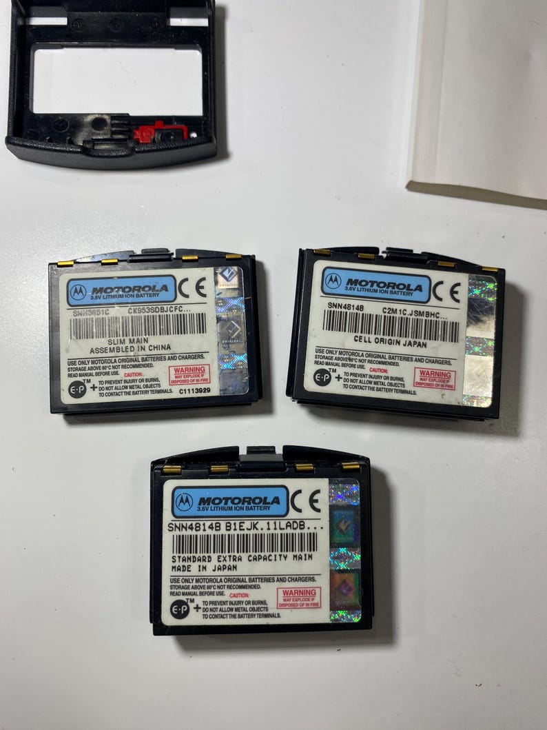 Puede incluir: Tres bater&iacute;as de iones de litio Motorola de 3,6V en color negro con etiquetas blancas que muestran especificaciones t&eacute;cnicas, textos de advertencia y pegatinas de seguridad hologr&aacute;ficas. Una bater&iacute;a est&aacute; marcada como Slim Main y otra como Standard Extra Capacity Main.