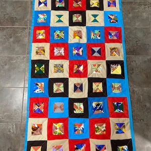 May include: A patchwork quilt with a colourful design. The quilt is made up of a grid of squares in red, blue, beige, and black. Each square contains a smaller, patterned fabric square. The quilt is edged with a bright blue fabric.
