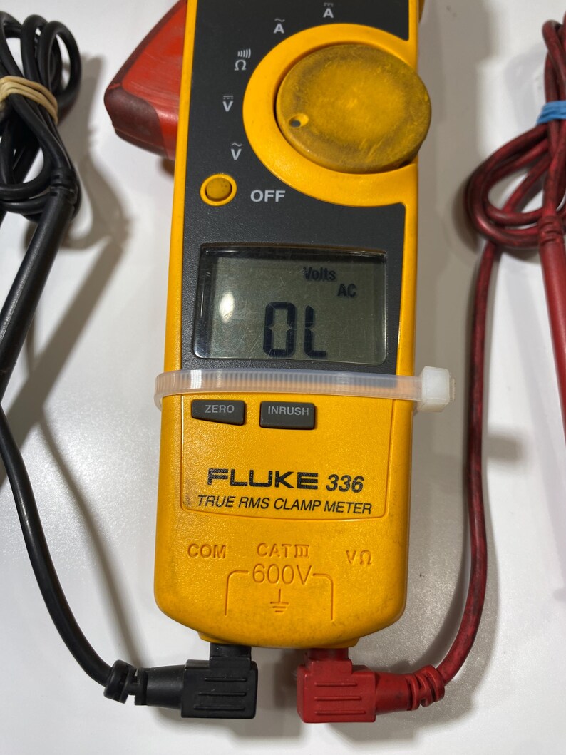 Pu&ograve; includere: Una pinza amperometrica Fluke 336 True RMS gialla con un display digitale che mostra OL e Volts AC. Il dispositivo presenta una grande manopola circolare, pulsanti funzione etichettati ZERO e INRUSH e puntali neri e rossi collegati alla base.