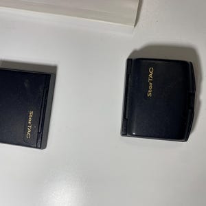 Puede incluir: Dos bater&iacute;as rectangulares negras para tel&eacute;fono m&oacute;vil de un dispositivo StarTAC. Cada unidad presenta un acabado mate liso con el nombre de la marca StarTAC impreso en letras doradas en la parte frontal. Los componentes est&aacute;n colocados lado a lado.